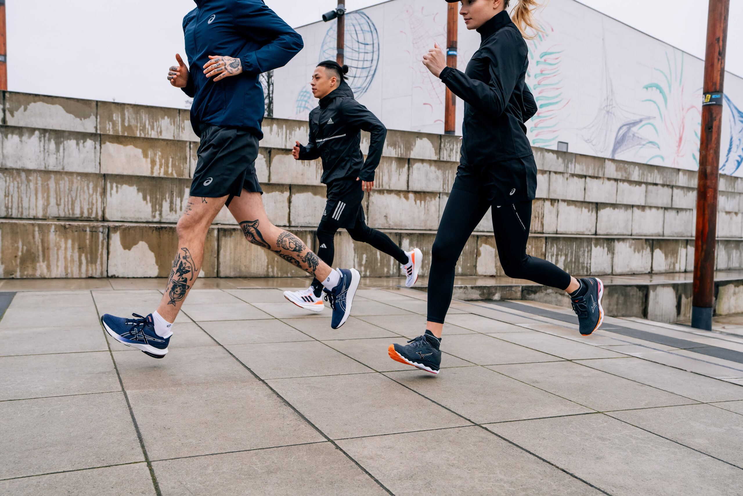 trīs skrējēji skrien pa bruģi ar adidas, asics un mizuno apaviem
