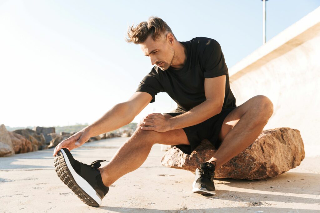 Mężczyzna rozciągający się przed porannym joggingiem na plaży w czarnej koszulce, spodenkach i sportowych butach męskich