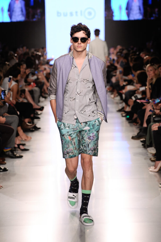 Model ubrany w letnią stylizację: wzorzysta koszula i shorty, fioletowa bomberka oraz białe klapki Puma zestawione z wzorzystymi skarpetkami